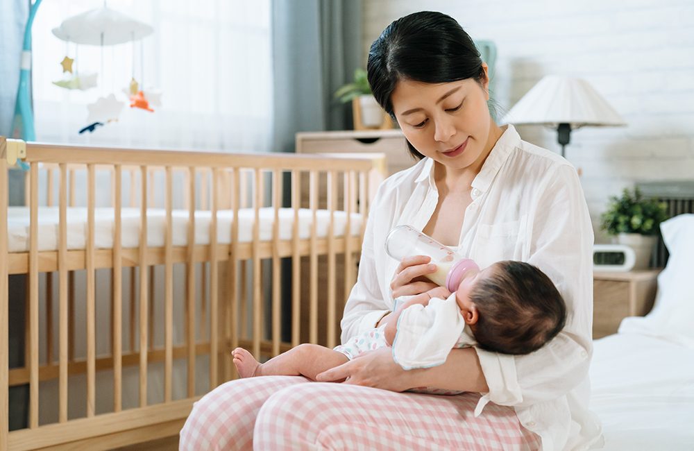 portrait japanese woman sitting at bedside providing infant formula to her baby. asian mom holding her child in arms and feeding her with a loving look. mother's day concept.
